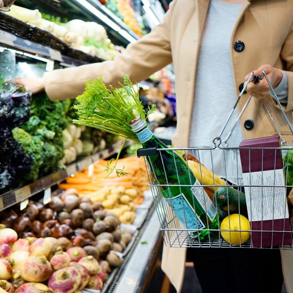 A person shopping with a basket full of groceries, including celery and a bottle of water