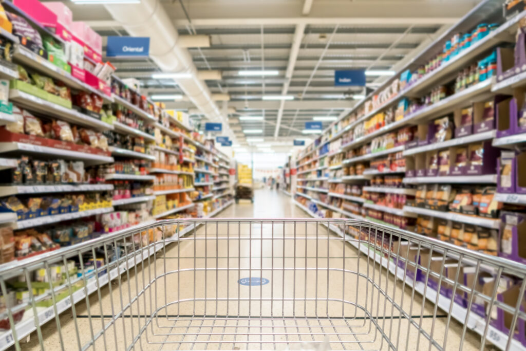 image shows a shopping trolley in a supermarket, pointing down an aisle with food on either side