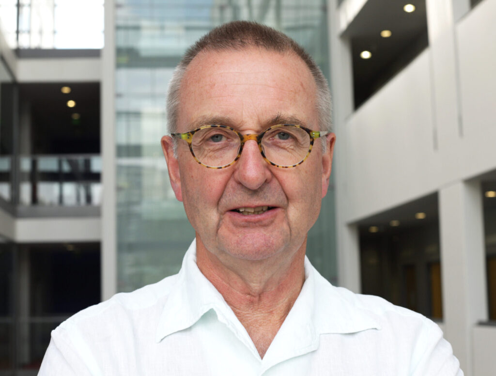 Image is a headshot of Tim Lang. He is a wearing glasses and a white shirt and standing in front of a university building.